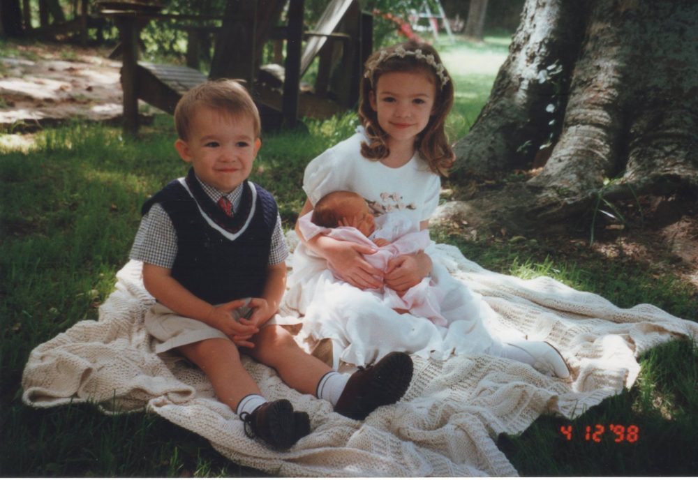 Two young children, a boy and a girl, sitting on a picnic blanket holding a baby; the date 4/12/98 in the corner.