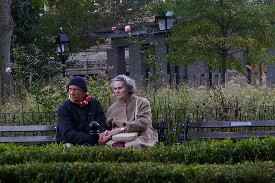 Color photograph: older couple sits and holds hands on a park bench, both looking toward an unseen point
