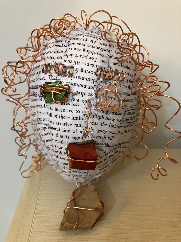 Straight-view of a papier mache head with text for skin; copper-wire hair, nose, and, eybrows; copper-wire affixes a green glass as a right eye, clear glass as a left eye, and a red brick mouth; stone base.
