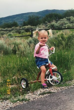 A young girl with pigtails and a baby pink shirt sits on a bike on the side of the road. 