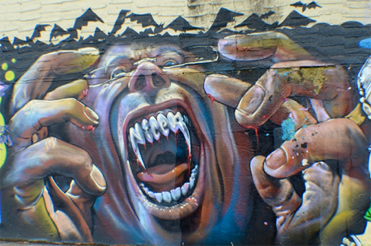 Photograph of graffiti mural depicting a screaming face with vampire teeth, and fleshy hands raised in a claw-like gesture, bats rising in silhouette in background