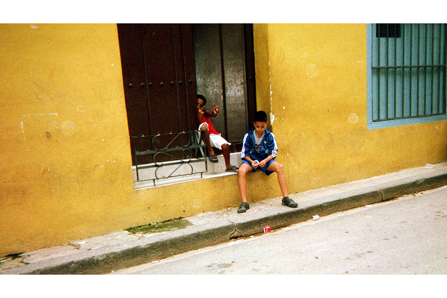 Two young boys sit in the open window of a yellow building; one shoots peace signs toward the camera, the other plays with something in his hands.