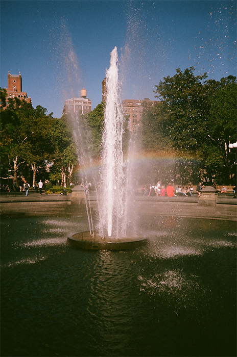Photograph of a jet of water from a park fountain, partial rainbow in foreground, trees, buildings, and deep blue sky in background.