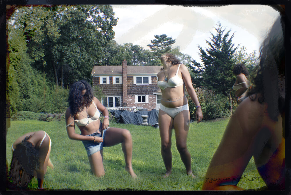 Multiple of the same figure in different stages of disrobing in a lush green yard before an old house. Their face is not shown.