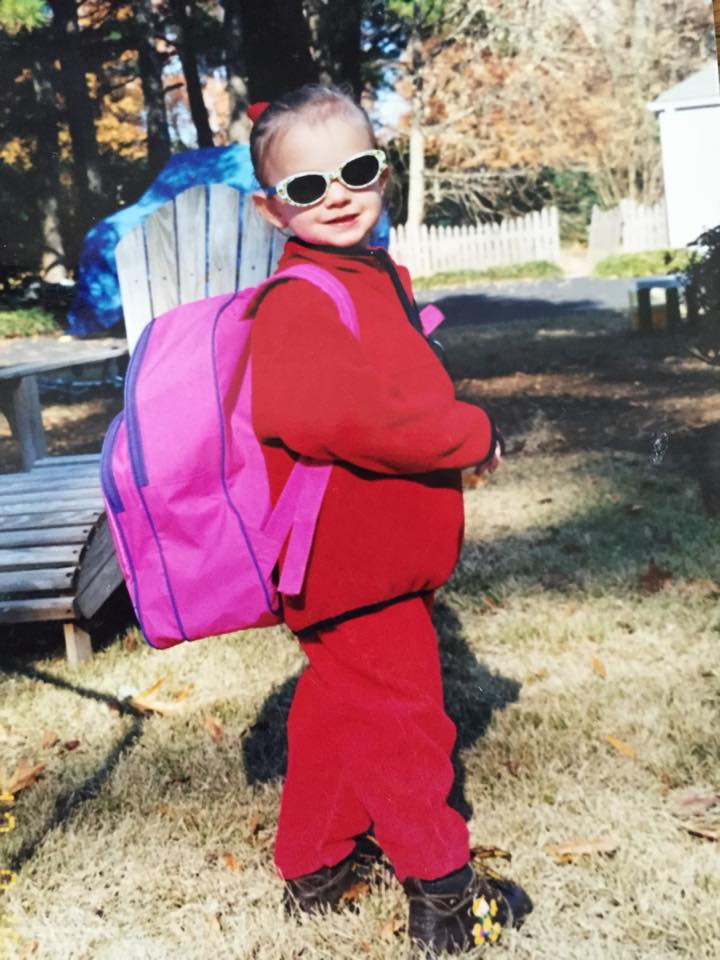 A young girl in a red fleece and sweatpants and sunglasses, posing with a big pink backpack.