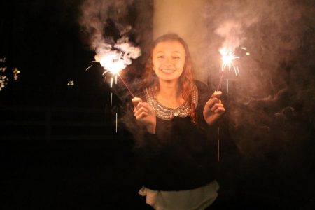 A girl smiles for the camera, holding two sparklers around her face. 
