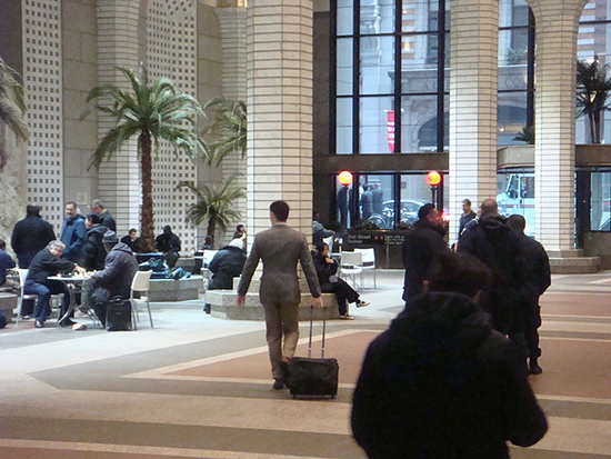 The hustle and bustle of 60 Wall street: people walking around, a man rolling a suitcase, people sitting at tables. 