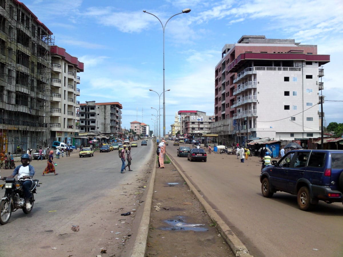 Photograph showing a street, its median at the center, sparse pedestrians and cars on the sand-colored road, mid-rise buildings on both sides