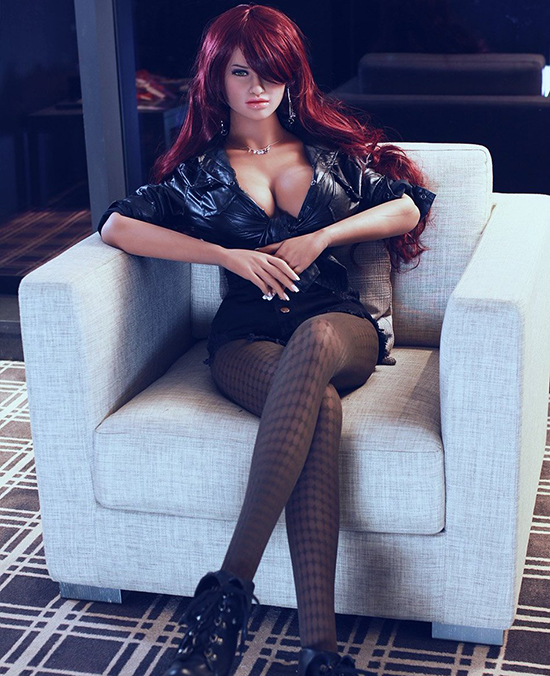 Photograph of life-size doll, with long read hair and ample cleavage, seated in a beige armchair