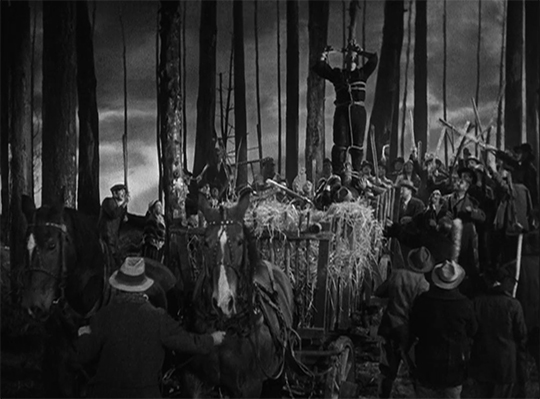 Black and white film still still in which a mob carries the Creation, bound to a pole, through a forest; those in the background wield sticks In the scene; in the foreground, a man leads a two-horse cart