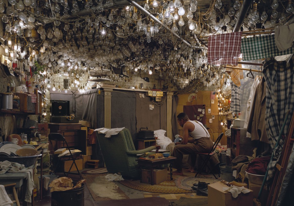 "Invisible Man" photograph of a man sitting hunched over in a room filled with light bulbs. 