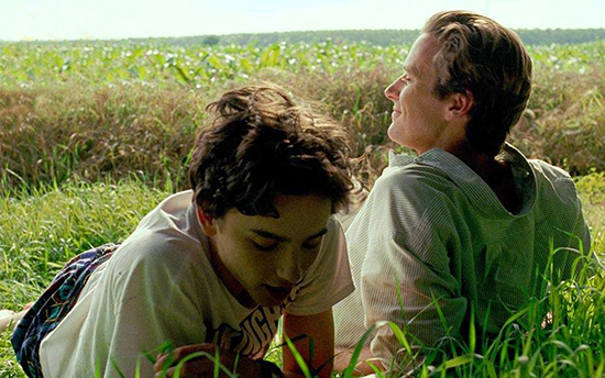 Two men lay in the grass, one sitting and looking out at the view, the other laying on his stomach, looking down.