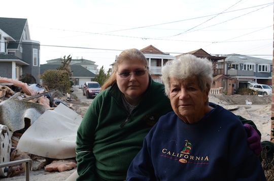 Two women, looking into the camera, posed in front of a neighborhood and its wreckage.