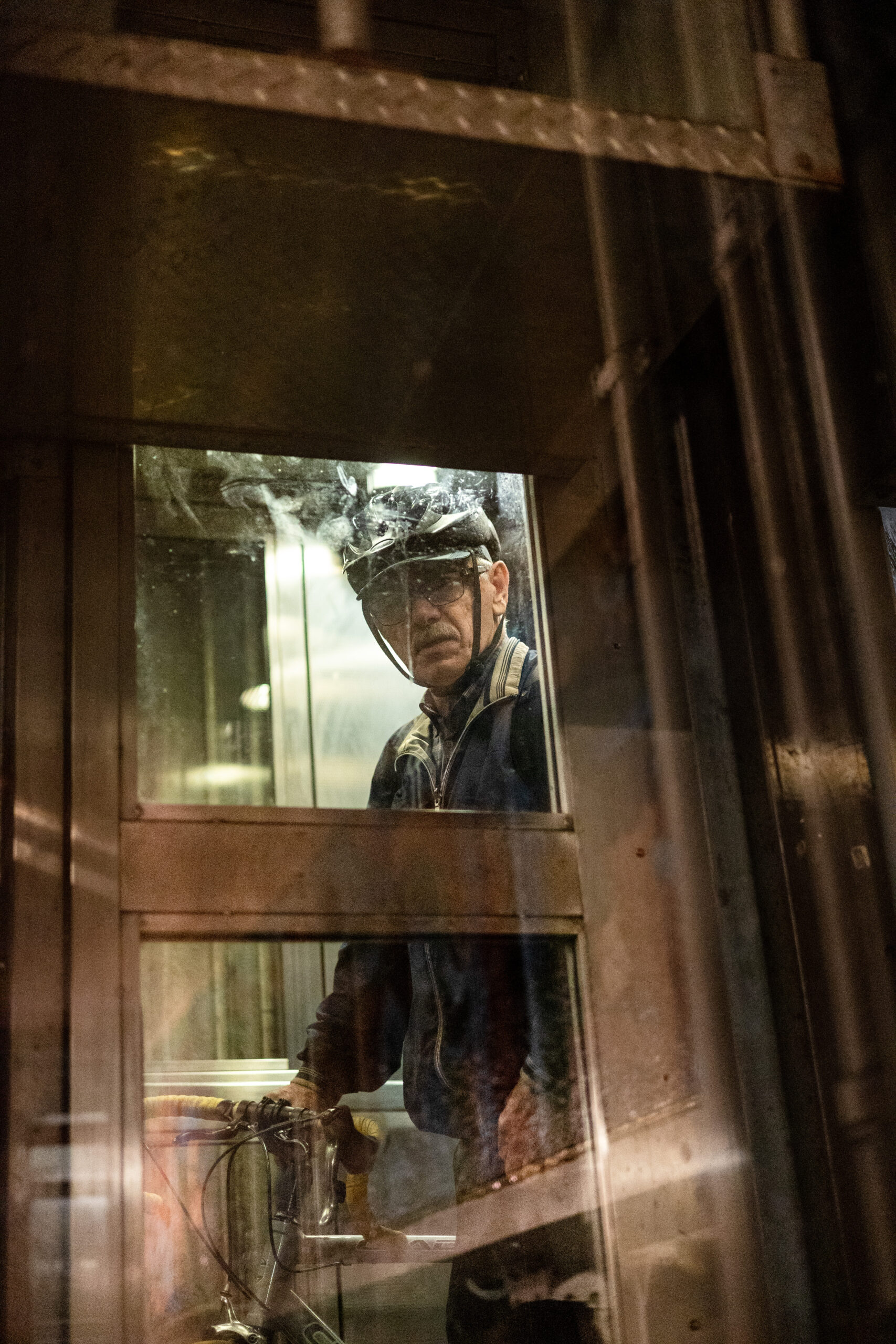 Trapped behind a grid of glass and metal a man looks down, directly into the lens with slight confusion. Luminescent light shines from abroad and illuminates the smudges and grim on the glass. He is wearing a biking helmet and holding onto a bike.