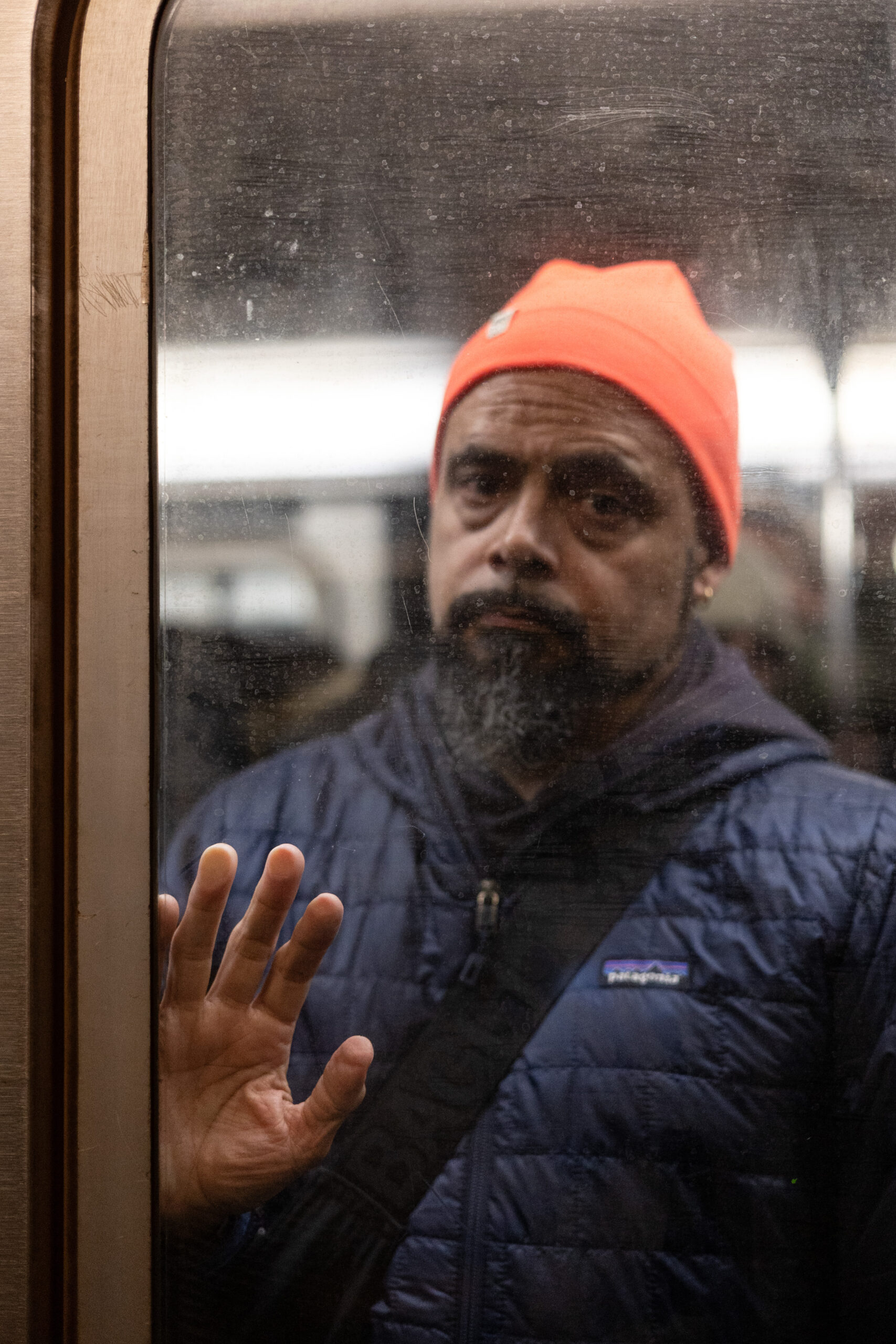 A bearded man presses his fingertips against the glass of the subway door. He is making eye contact and his eyebrows are raised though there is no expression on his face. He is wearing a bright orange toque and a dark blue jacket. The image is framed by stripes of metal and rubber going down the left side of the image. 