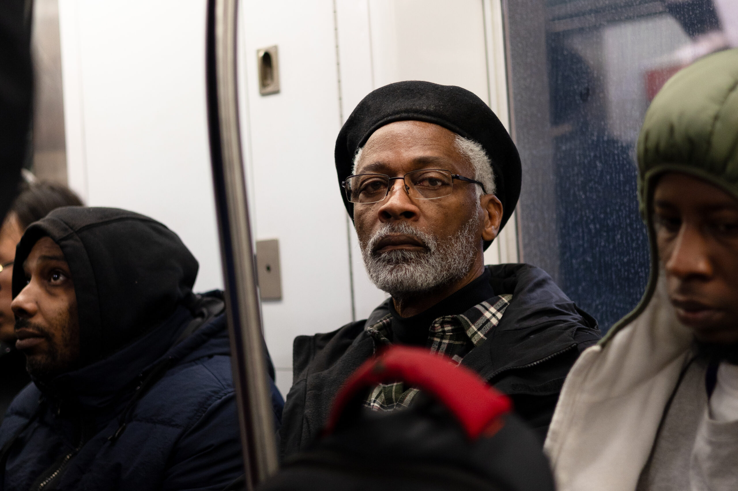 Seated in a subway car, a man stares directly at the camera with no expression on his face. His white beard and hair contrasts with his dark features. He is wearing a beret and glasses.