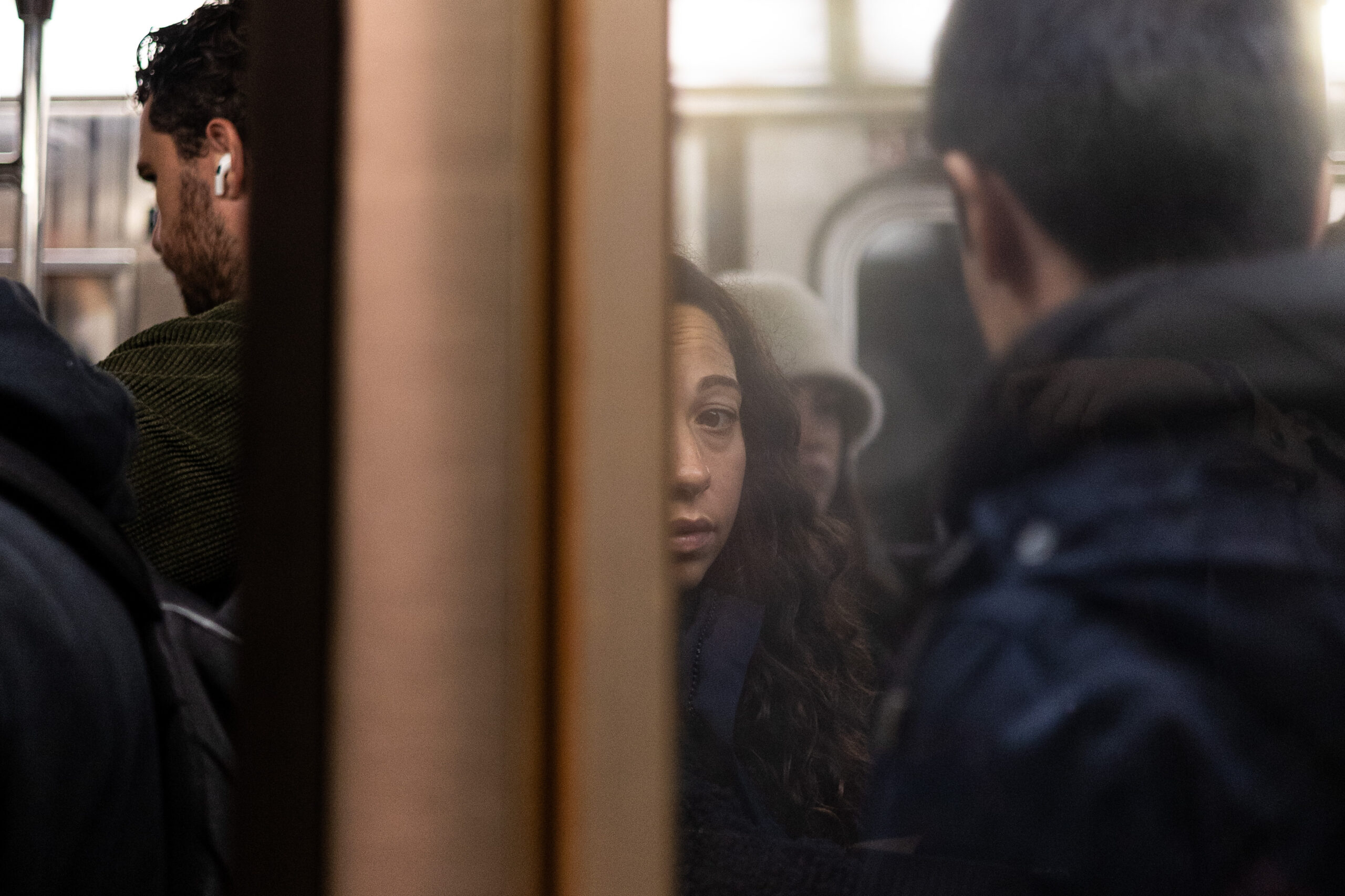 A woman looks directly into the camera as she stands expressionless in a crowded subway car. Exactly half of her face is covered by the opening subway door. On both sides of her are men with their backs turned towards the door.
