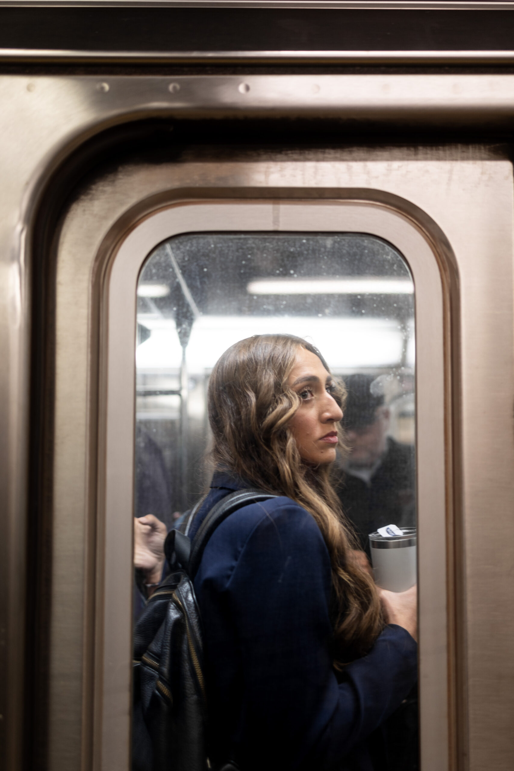 A woman looks into the distance from inside a subway car as she waits for the subway doors to open. She is holding an yeti and wearing a blue blazer. Her profile is framed by the subway door and illuminated by the luminescent lights from above.