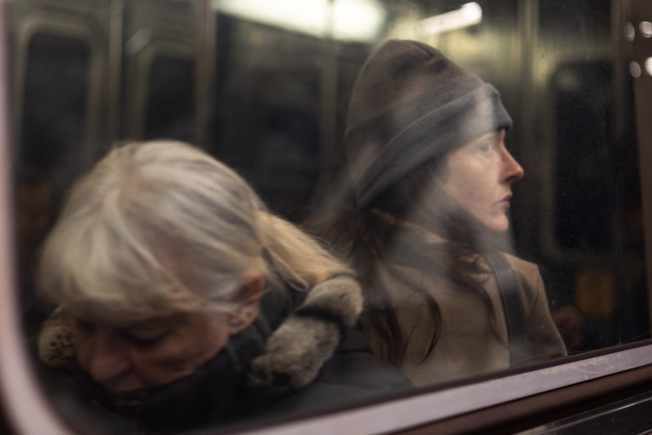 A woman stares into the abyss from inside a subway car. Her face is illuminated by a triangle of light cast from the reflection on the car’s window. Her back is towards the camera and only her profile can be seen. She is wearing a grey toque and a brown wool coat, which is a similar colour to her hair.  An older woman sitting closer to the camera looks down at her lap. The women sit back to back and neither are in focus. They are framed by strokes of metal.