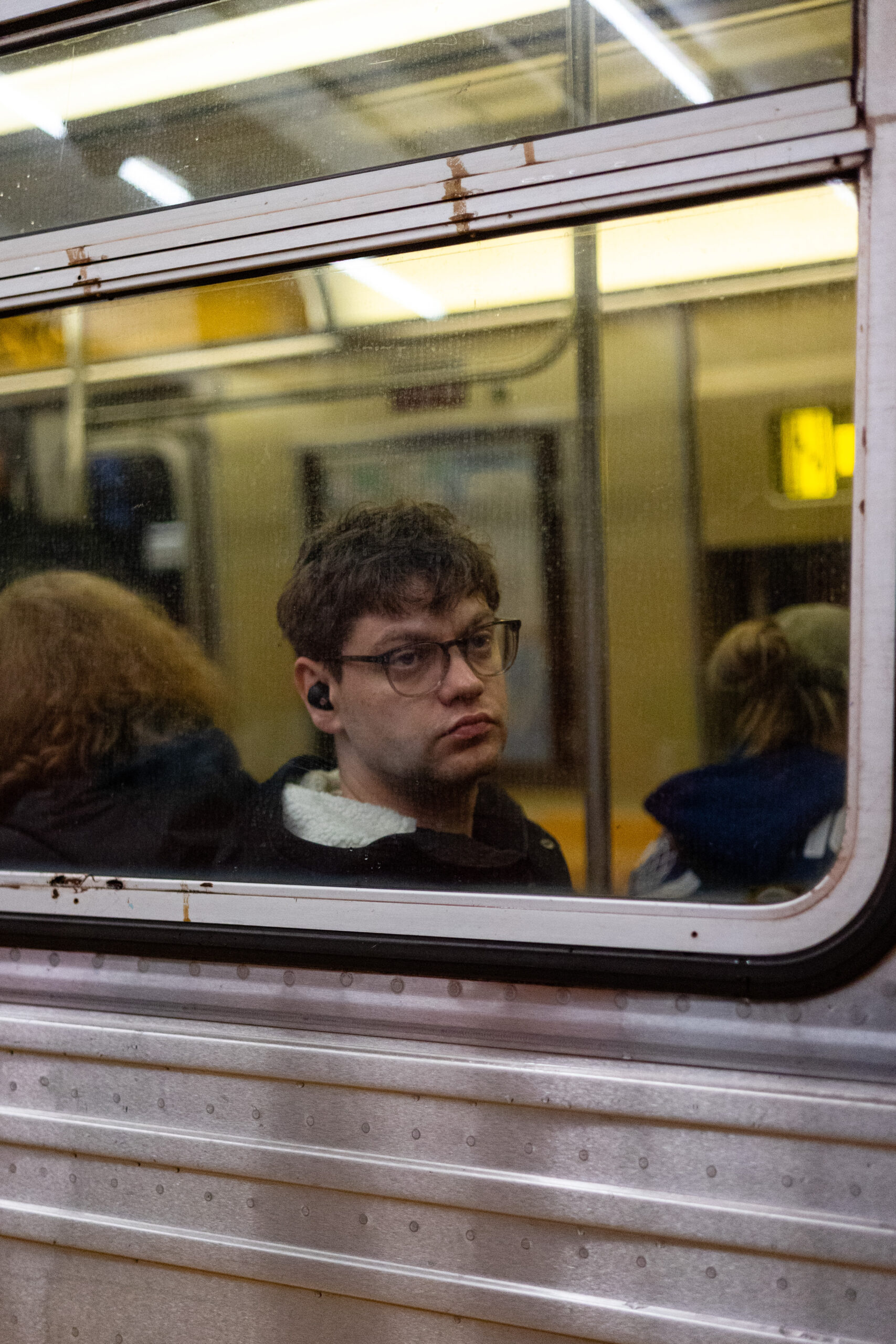 A man looks out the subway window seemingly at nothing. His face and eyes are expressionless. Two woman sit on either side of him, their back towards the camera. Skid marks can be seen on the dirty window and there are stripes of light reflection from the luminescent lights above. He is framed by the cool silver of the subway car which contrasts with the warm interior.