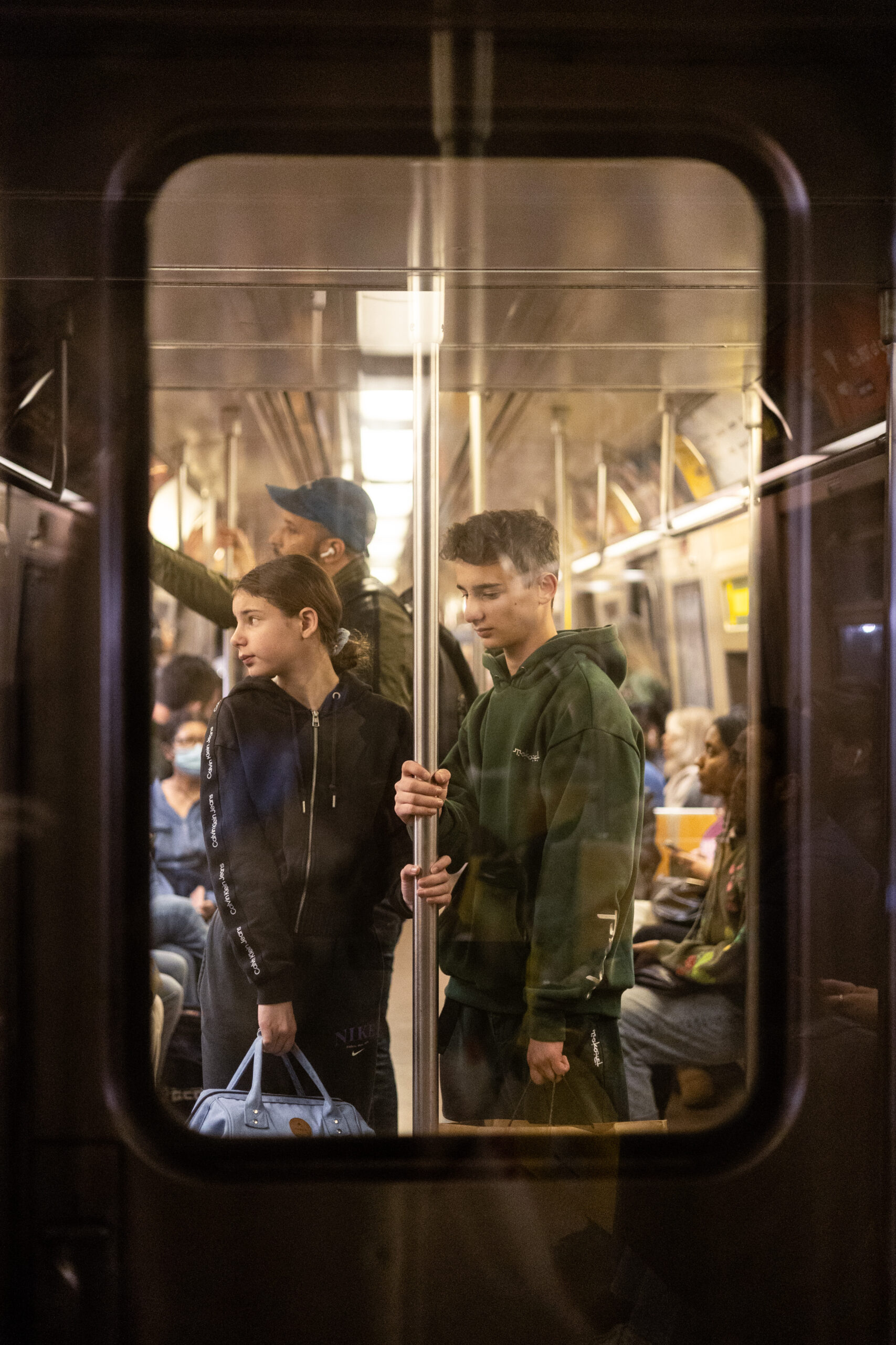 A young woman and a young man stand at the front of the next subway car sharing a pole. While she looks to the left at something in the distance, he looks down at her feet. They are framed by the window at the end of the subway. The reflection off two panes of glass casts linear stripes of light which continue out of frame. The two panes of glass also cast a reflection of the pole which extends out of the primary frame. 