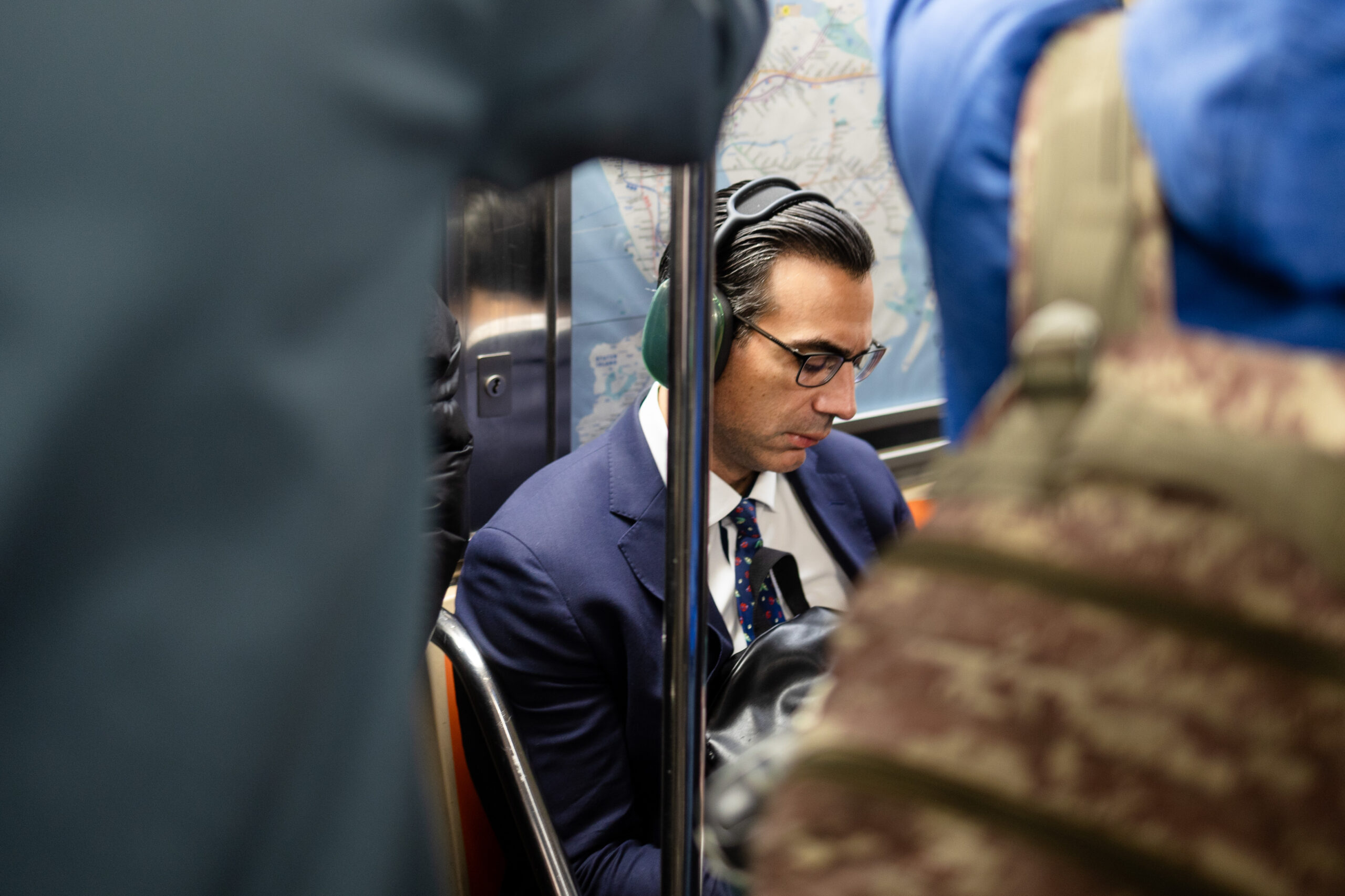 A man in a blue suit and tie is seated in a crowded subway car looking down at something in his hand which is blocked by a green backpack. He is framed by the backs of people standing in the foreground, both also in various shades of blue. His hair is jet black and slicked back. He is wearing a pair of green headphones and glasses. Behind him is a blue map which contrasts with the hint of orange on either side of him.