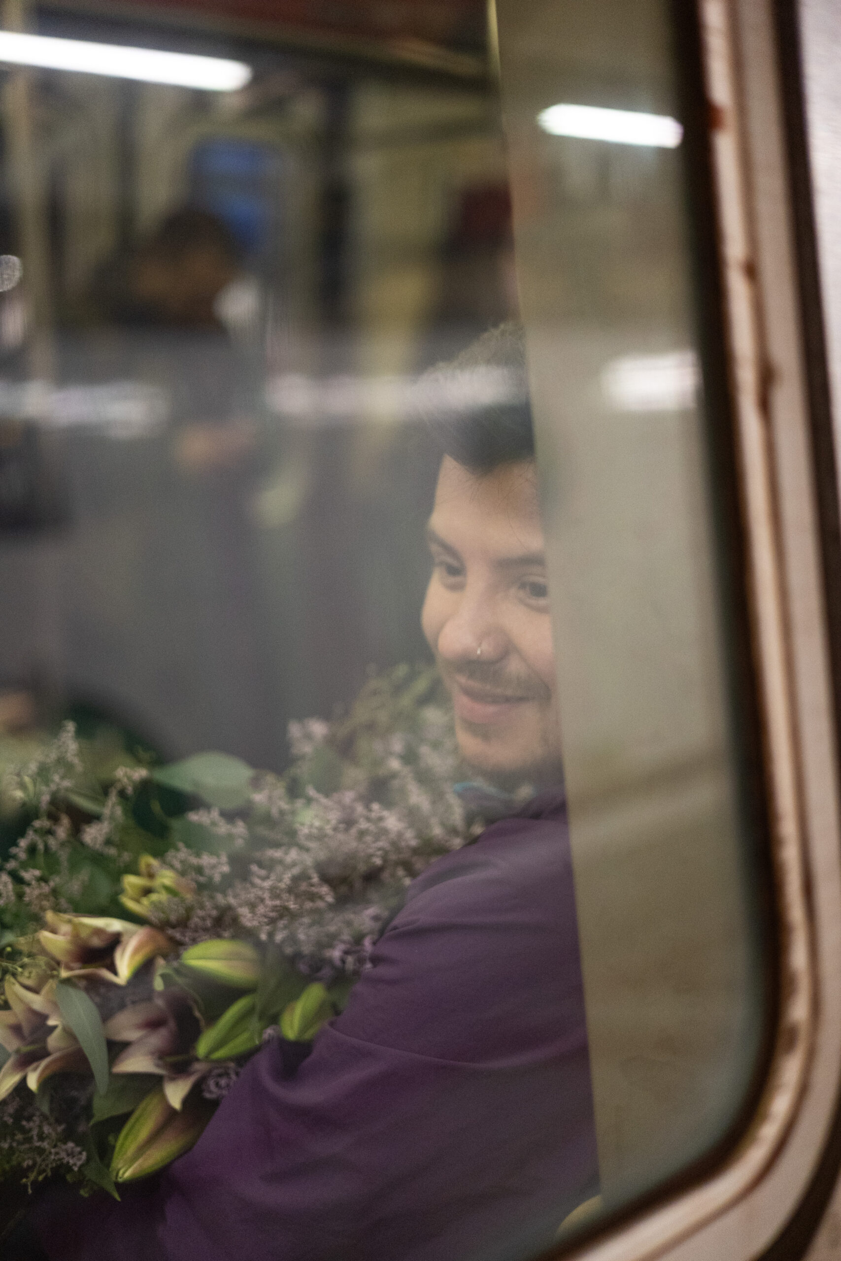  A man holding a large bouquet of lilies sits with his back towards a subway window. He looks at something out of frame with a look of quiet contentment. Only part of his face can be seen. He is wearing a warm purple dress shirt which is of a similar color to the flowers in his arms. The smudged reflections of people in the subway are distorted by stripes of light cast by the luminescent lights above. A stroke of metal frames the image.