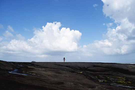Photograph of a wide, flat expanse of lava rocks under a blue sky with cumulus clouds; in the distance but at the center of the frame stands a person 