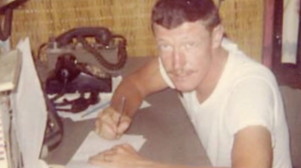 A man in a white undershirt sits at a small desk writing a letter; he glanced at the camera. 