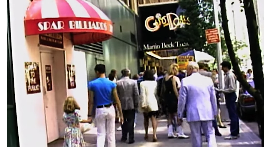 Photograph of street scene; a crowd including Rafsky and young daughter are seen from the back, in summer dress, walking toward The Martin Beck Theatre, where the marquee announces "Guys and Dolls"