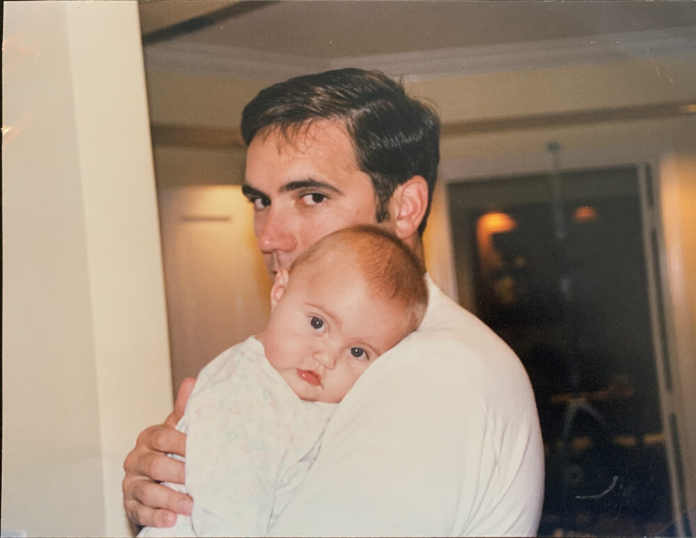 Photograph of a man in profile holding a baby, whose head rests on his shoulder to face the camera straight on.