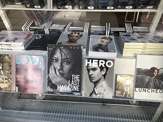 Photograph through a bookstore window showing a shelf display of fashion magazines in two rows