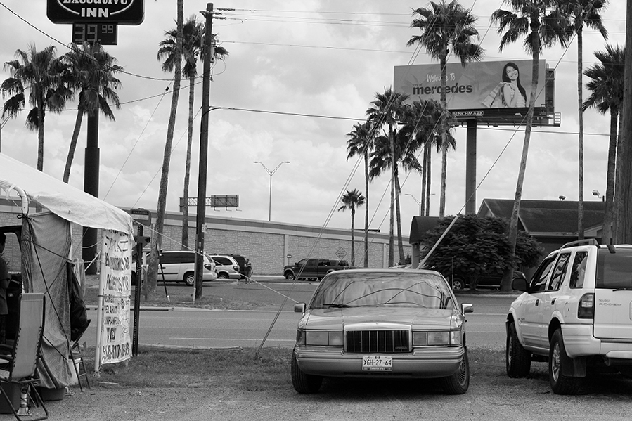 A view from a parking lot: a couple cars, a tent to the left, and a billboard by the highway (in black and white). 