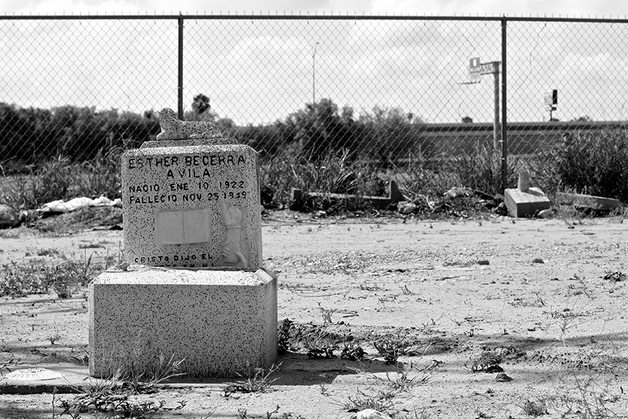A small stone on a dirt road in front of a chainlink fence, reading "Esther Becerra / A Vila / Nacio Ene 10 1922 / Fallecio Nov 25 19??"