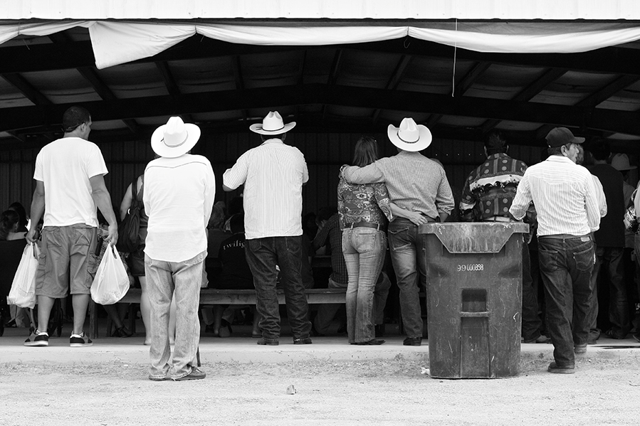 The backs of a crowd, mostly wearing cowboy hats, as the stand and sit watching an unseen stage (in black and white).