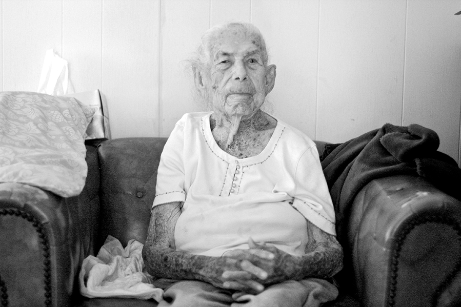 Portrait of an older woman sitting on a sofa, gazing toward the camera (in black and white). 