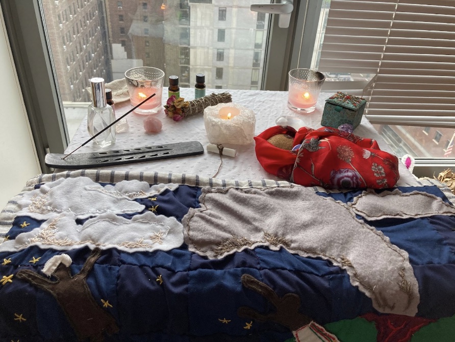 The finished quilt, incense, and lit candles sit on a table by a bright window. 