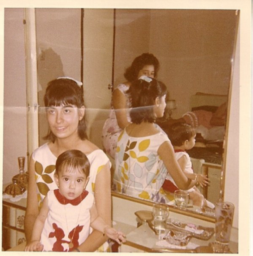A photograph of a child sitting on a young woman's lap, in front of a vanity table; the mirror shows the woman and child from the back, as well as another young woman, at whom the first young woman is looking