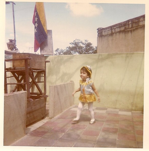 A toddler in a yellow and white dress, the Venezuelanlag in the background