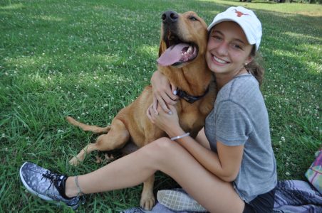 A young woman sits on green grass hugging her dog, wearing a Texas Longhorns cap. 