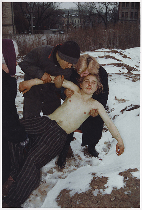 An intoxicated man with a naked torso is being held up by three other people. It is winter outside.