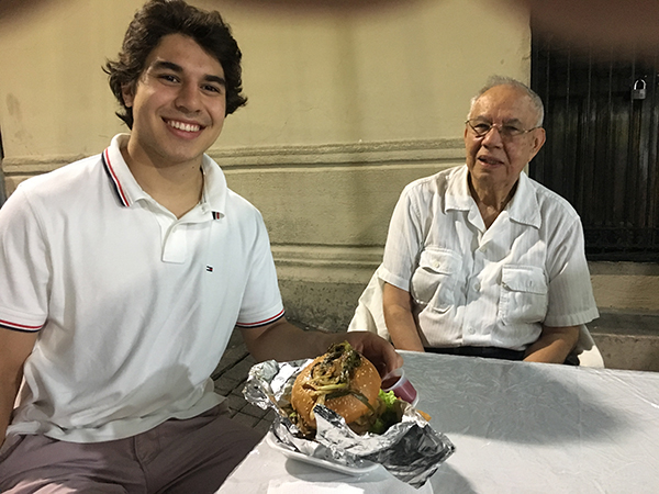 David Rosales and his grandfather sit around an outdoor table, both wearing big smiles. David has a large sandwich in front of him on foil.