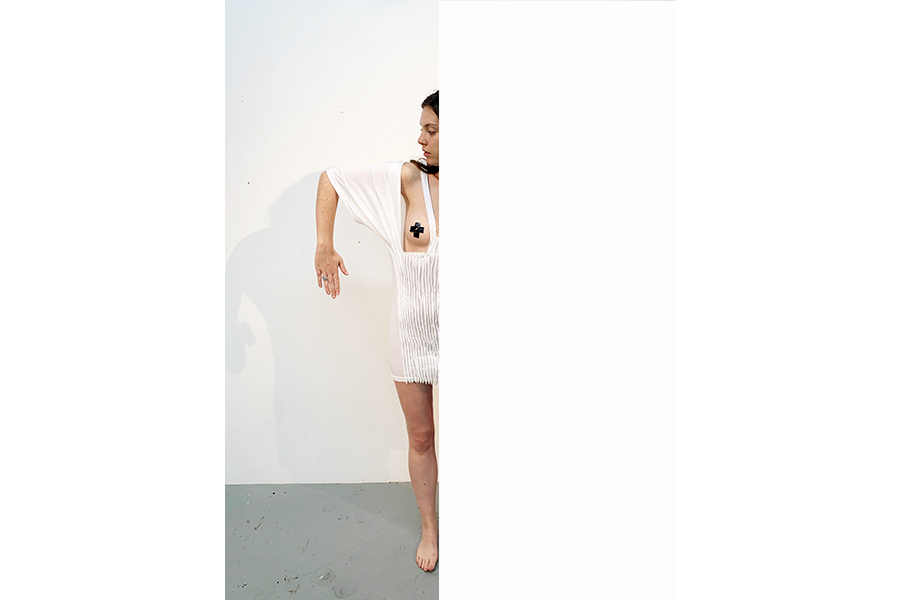 The model stands half obscured by a wall, showing the right side of the garment, right arm bent down at a right angle.