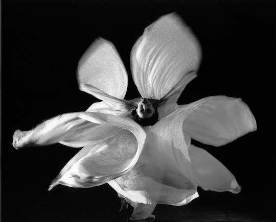 A black a white photo of Loie Fuller looking at the camera in a backbend while twirling white cloth into a symmetrical flower shape.