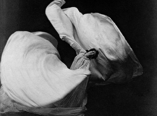 A black a white photo of Loie Fuller looking at the camera leaning towards the right while twirling white cloth into asymmetrical flower shape.