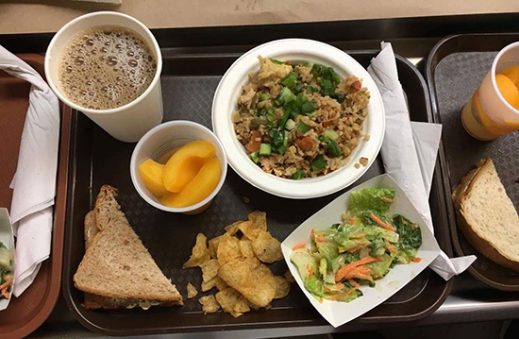 An overhead look at a lunch tray: a rice bowl, side salad, fruit cup, chips, bread, and coffee. 