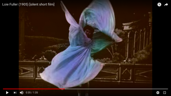 A recolored blue and purple photo of Loie Fuller looking at the camera leaning towards the right while twirling multicolored cloth into asymmetrical flower shape.