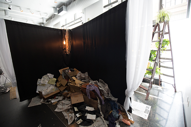 A corner encased in black sheets, with a pile of newspapers, cardboard boxes, and scraps of clothes. 