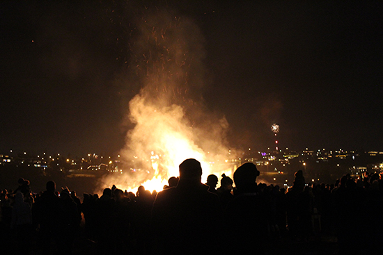 Figures in silhouette before a large fire. Nightime cityscape in distance 