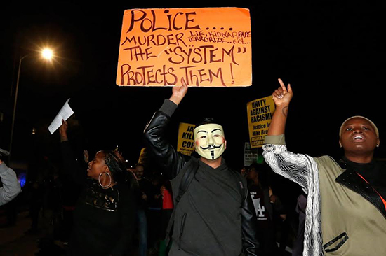 Protesters, one wearing a Guy Fawkes mask and carrying a sign that says "Police murder, lie, kidnap, rape, terrorize, etc... the "system" protects them!"